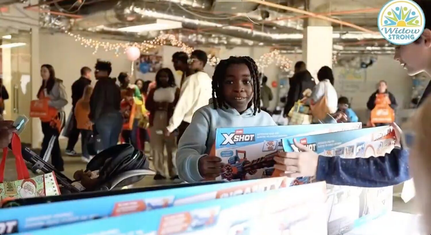 A smiling child in a light blue hoodie holds a large boxed toy at the gift tables while another family member reaches for a matching box