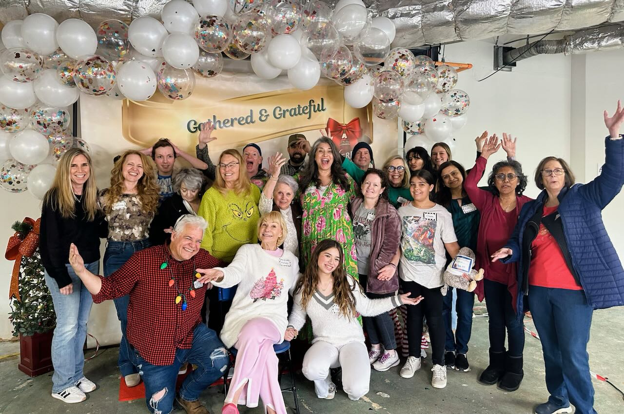 Family members and volunteers of all ages posing together beneath a balloon arch in front of a Gathered & Grateful backdrop