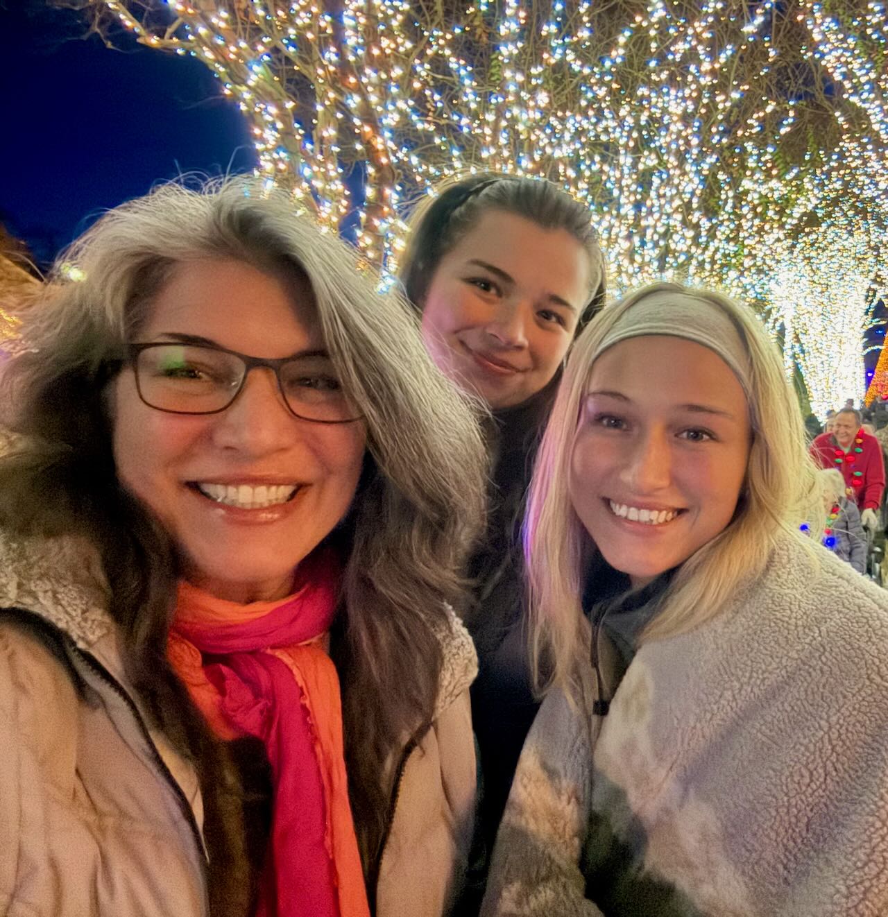 Mishael Porembski of Widow Strong, wearing glasses and a coral scarf, standing with two women beneath trees wrapped in holiday lights