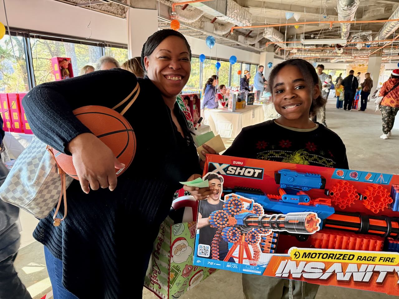 A smiling woman holding a basketball beside a young child proudly holding a large toy box at the gift gathering